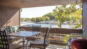 Une terrasse extérieure couverte, dotée d’une table et de chaises, d’un ventilateur de plafond, et offrant une vue sur un lac où l’on aperçoit des bateaux et des maisons au loin.