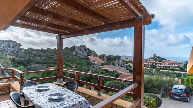 A covered outdoor seating area with a table and chairs, overlooking a scenic landscape with rocky hills and buildings. - Bella Vista (Trinità d'Agultu e Vignola)