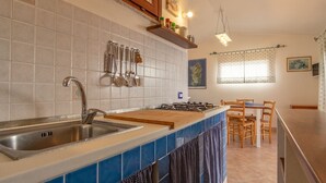 A kitchen with a sink, stove, and hanging utensils. There is a dining area with a table and chairs. - Bella Vista (Trinità d'Agultu e Vignola)