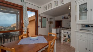 A dining area with a wooden table and chairs, a view through a window, and a kitchen with white cabinets. - Bella Vista (Trinità d'Agultu e Vignola)