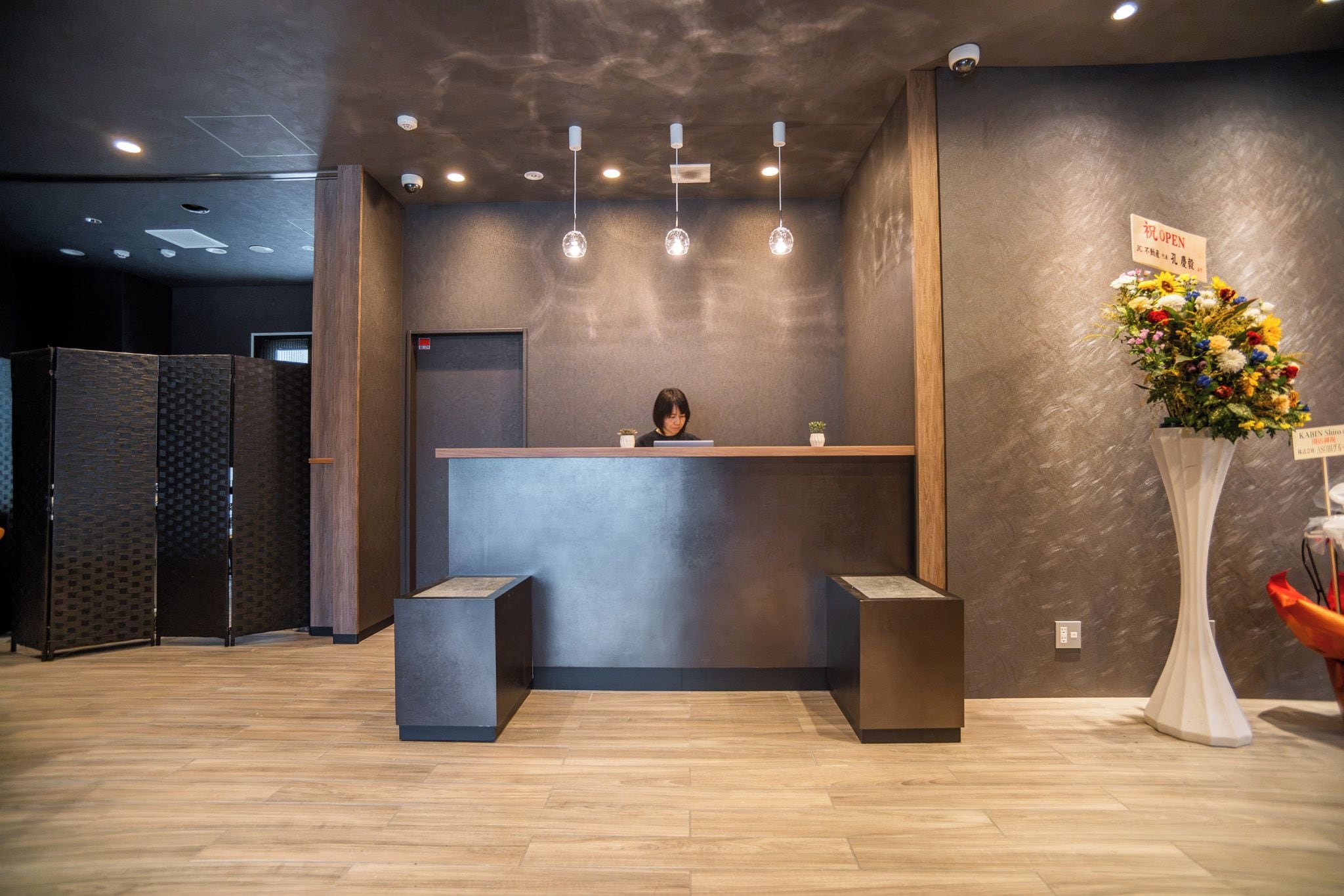 A reception desk with a person behind it, a modern light fixture above, and a vase with flowers to the right.