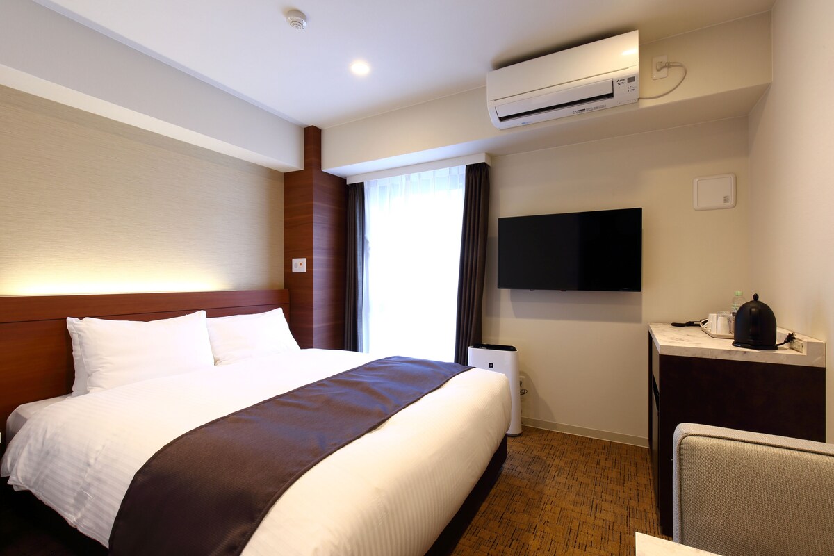 A hotel room with a large bed, an air conditioning unit, a desk with a chair, a flat-screen TV, and a window with curtains.