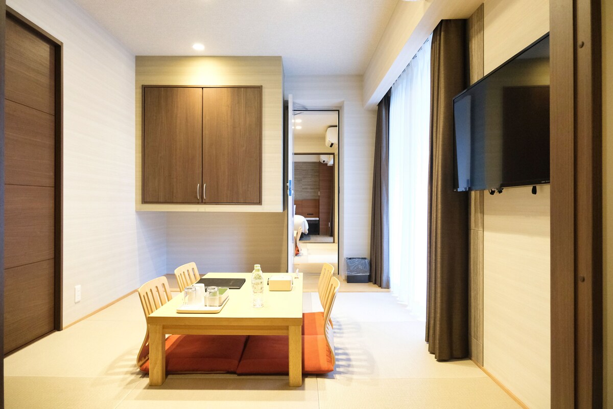 A modern hotel room with a dining area featuring a wooden table, chairs, and a sofa. A television is mounted on the wall, and a wardrobe is visible.