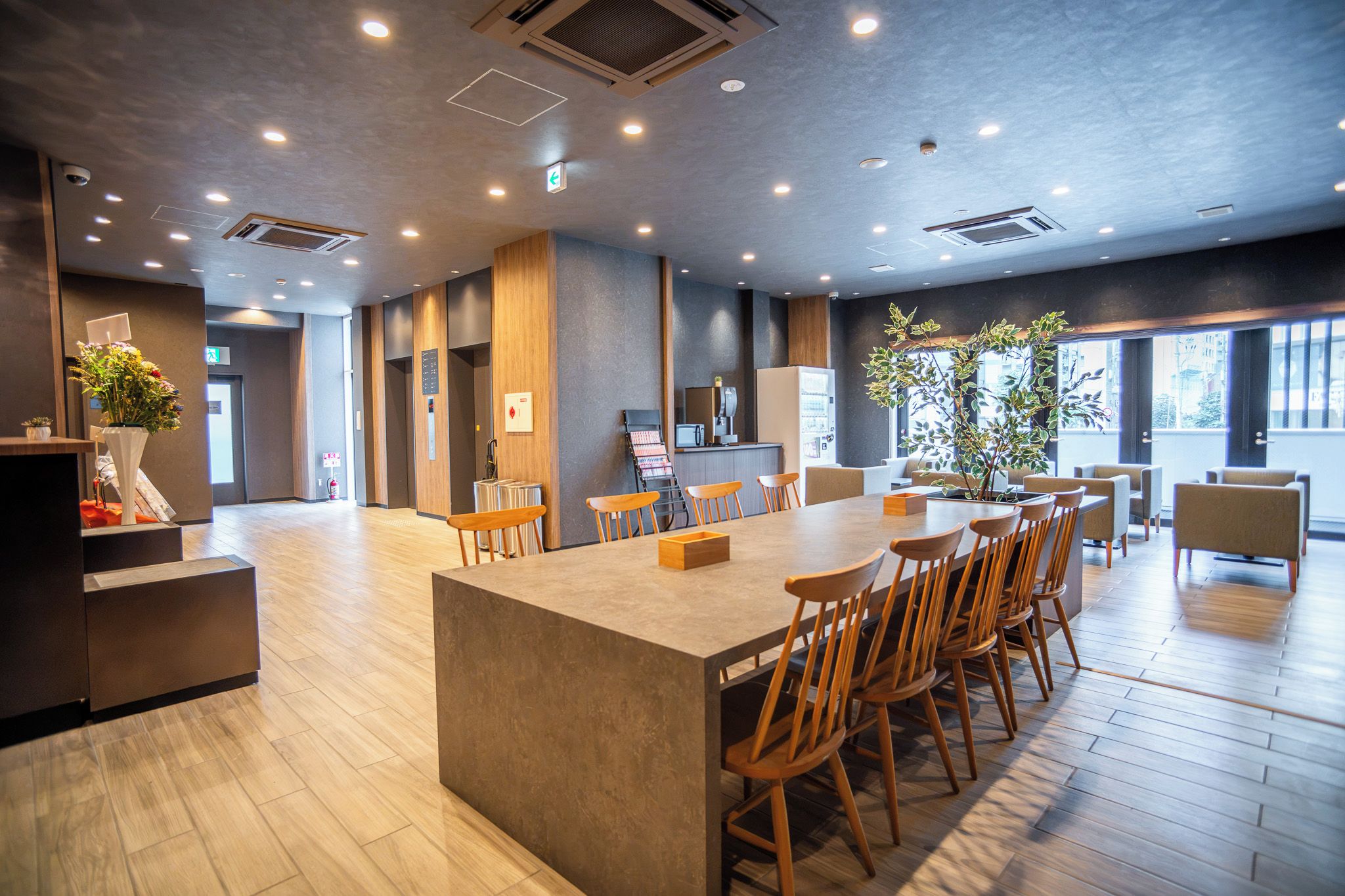 Un moderno vestíbulo de hotel con una mesa central tipo isla, sillas de madera, un mostrador de recepción y un ventanal con vistas a la ciudad.