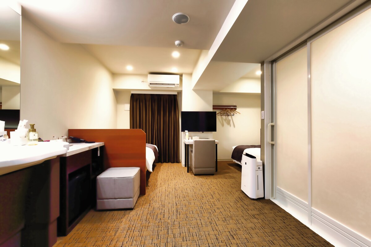 A hotel room with a bed, a desk, a chair, a small table, an air conditioning unit, a television, and a wardrobe.