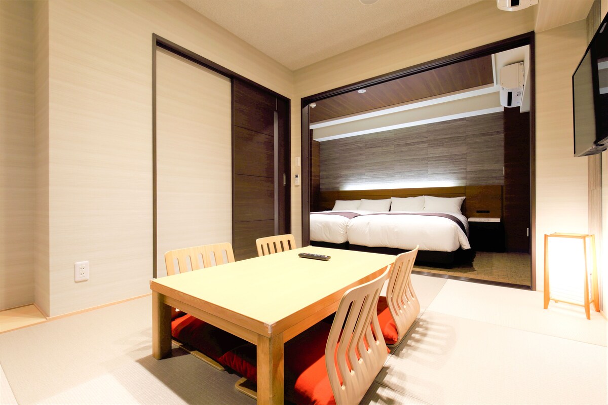A hotel room with a wooden dining table, chairs, a bed, and a TV.
