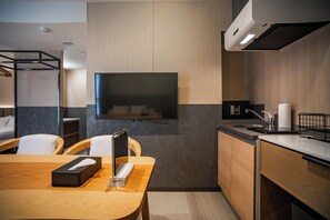 A modern hotel room with a wooden dining table, a flat-screen TV mounted on the wall, a small kitchen area with a sink and a safe, and a bed with a headboard. - Kabin Shiro - Regale Collection (Osaka)