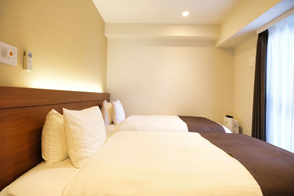 A hotel room with two beds, a wooden headboard, a bedside table, and a window with curtains.
