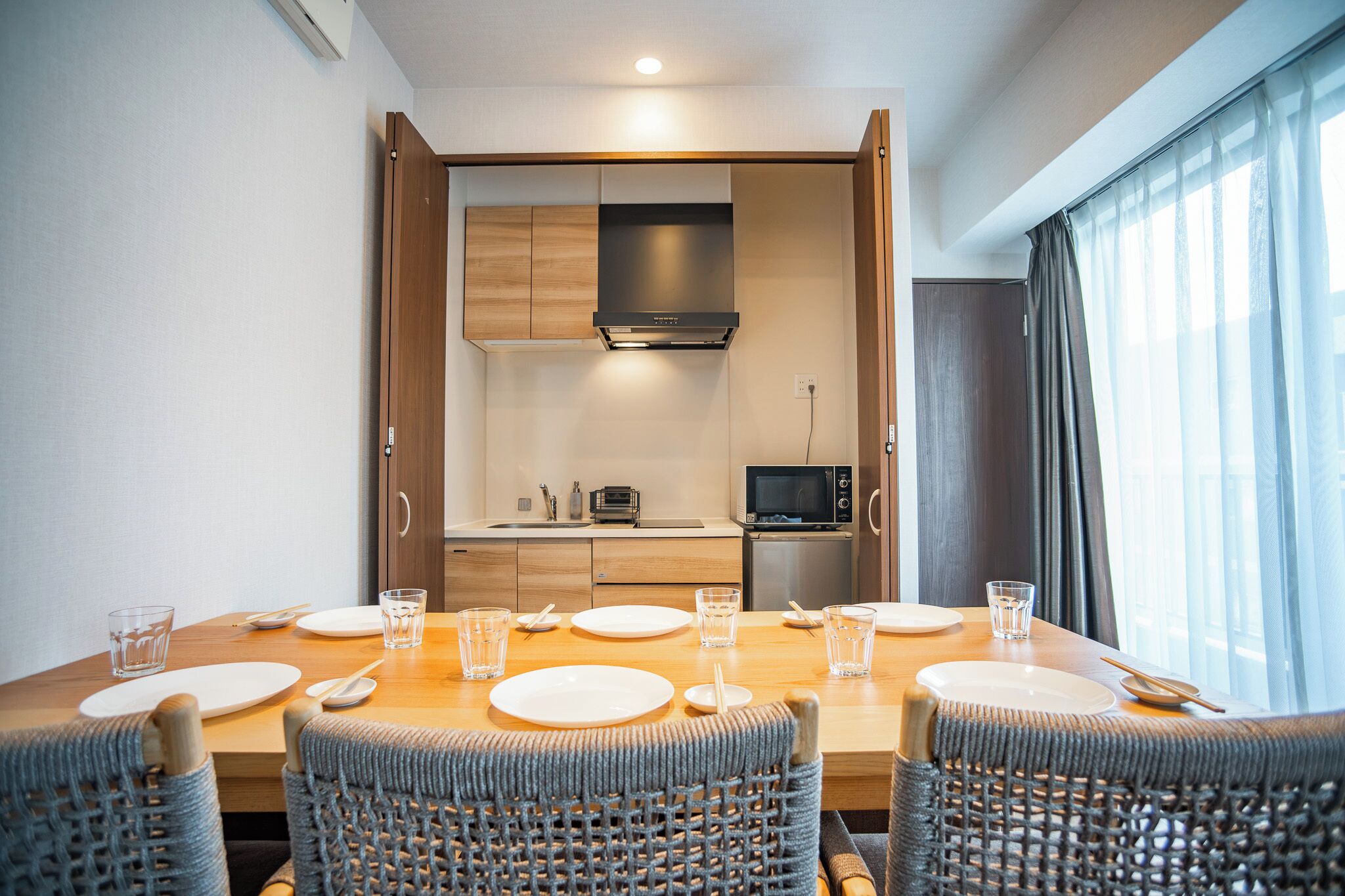 A dining area with a table set for four, a kitchen in the background, and a window with curtains.