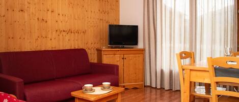 A room with a wooden panel wall, a red sofa, a wooden table with two cups, a television on a wooden cabinet, and a dining table with chairs.