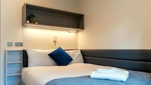 Un dormitorio ordenado con cama, mesita de noche, estante con plantas y una lámpara fijada a la pared.