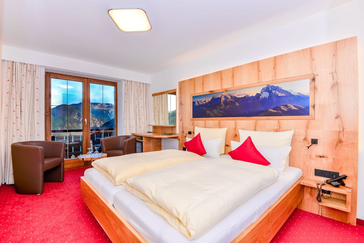 Ein Hotelzimmer mit einem großen Bett, zwei Stühlen, einem Schreibtisch und einem großen Fenster mit Blick auf die Berge.