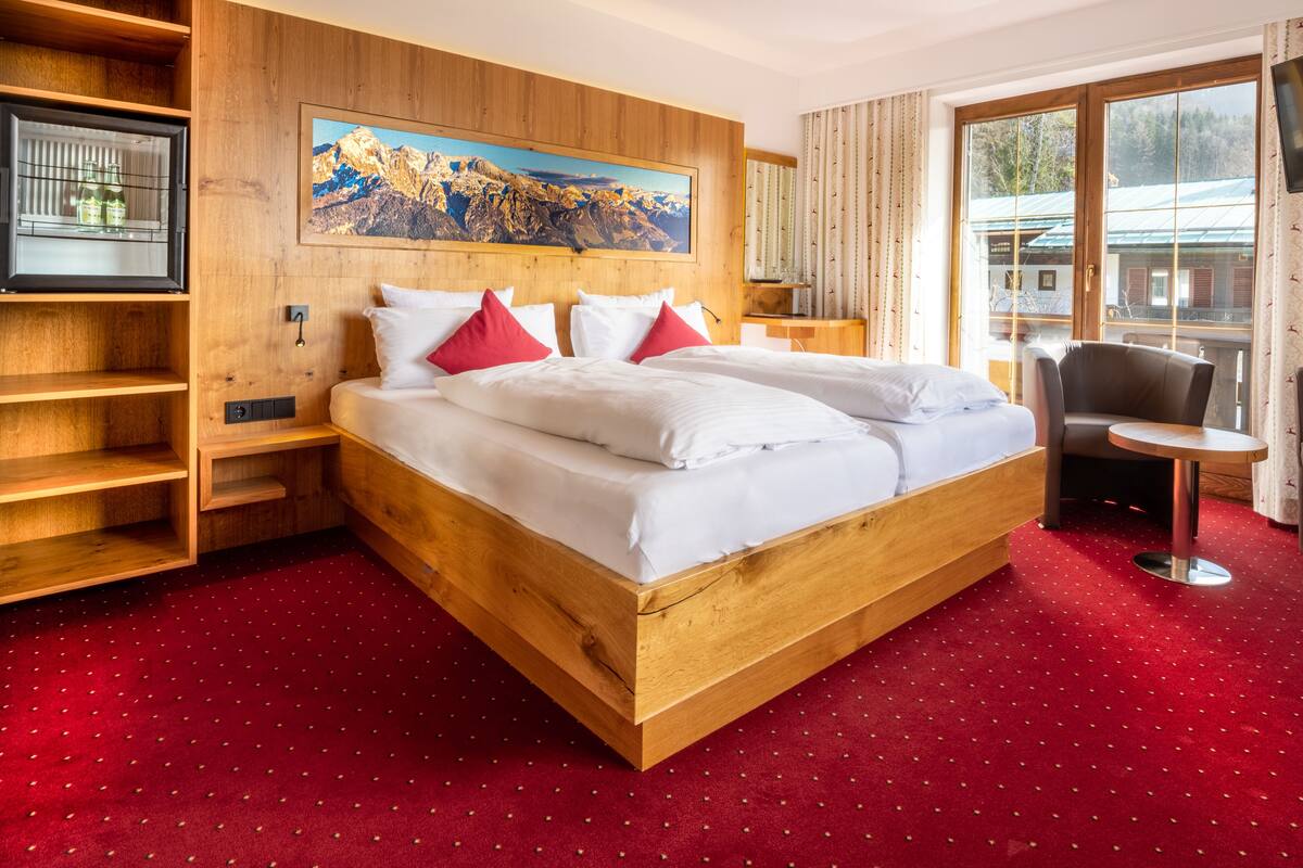 Ein Hotelzimmer mit einem großen Bett, einem Holzkopfteil, einem roten Teppich, einem Gemälde von Bergen und Blick auf ein Gebäude durch das Fenster.
