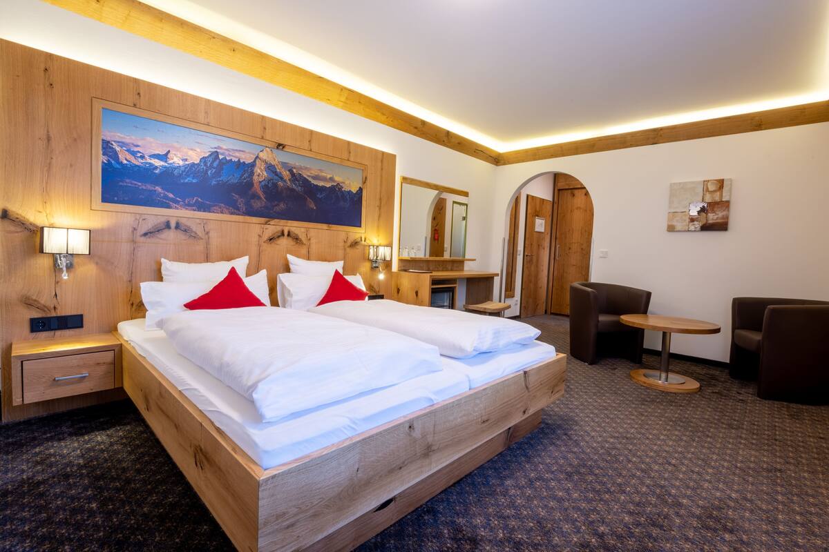 Ein Hotelzimmer mit einem hölzernen Bett, einem Bergpanorama, einem Schreibtisch, einem Sessel und einem kleinen Tisch.