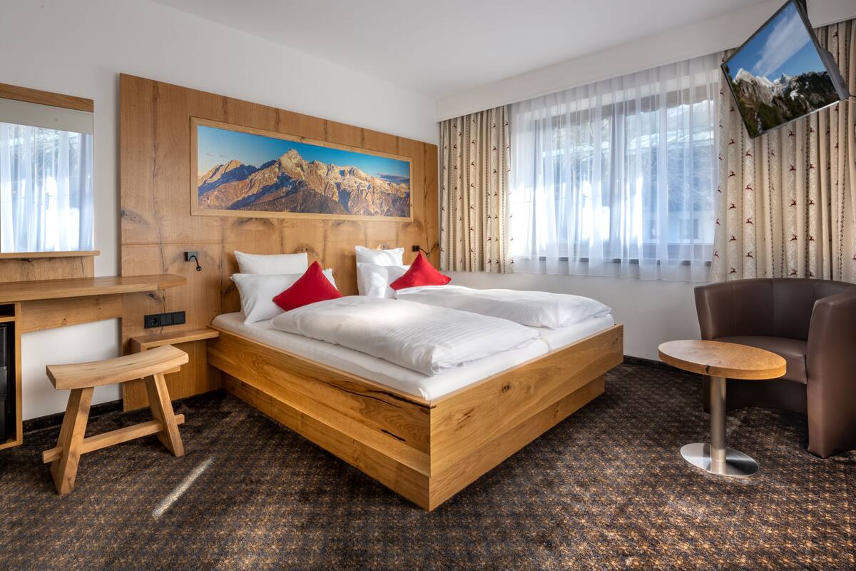 Ein modernes Hotelzimmer mit einem hölzernen Bett, einem Schreibtisch, einem Sessel und einem großen Fenster mit Vorhängen.