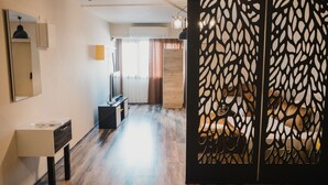 A modern hotel room with a wooden floor, a bed with a headboard, a desk with a chair, a mirror, and a decorative partition.
