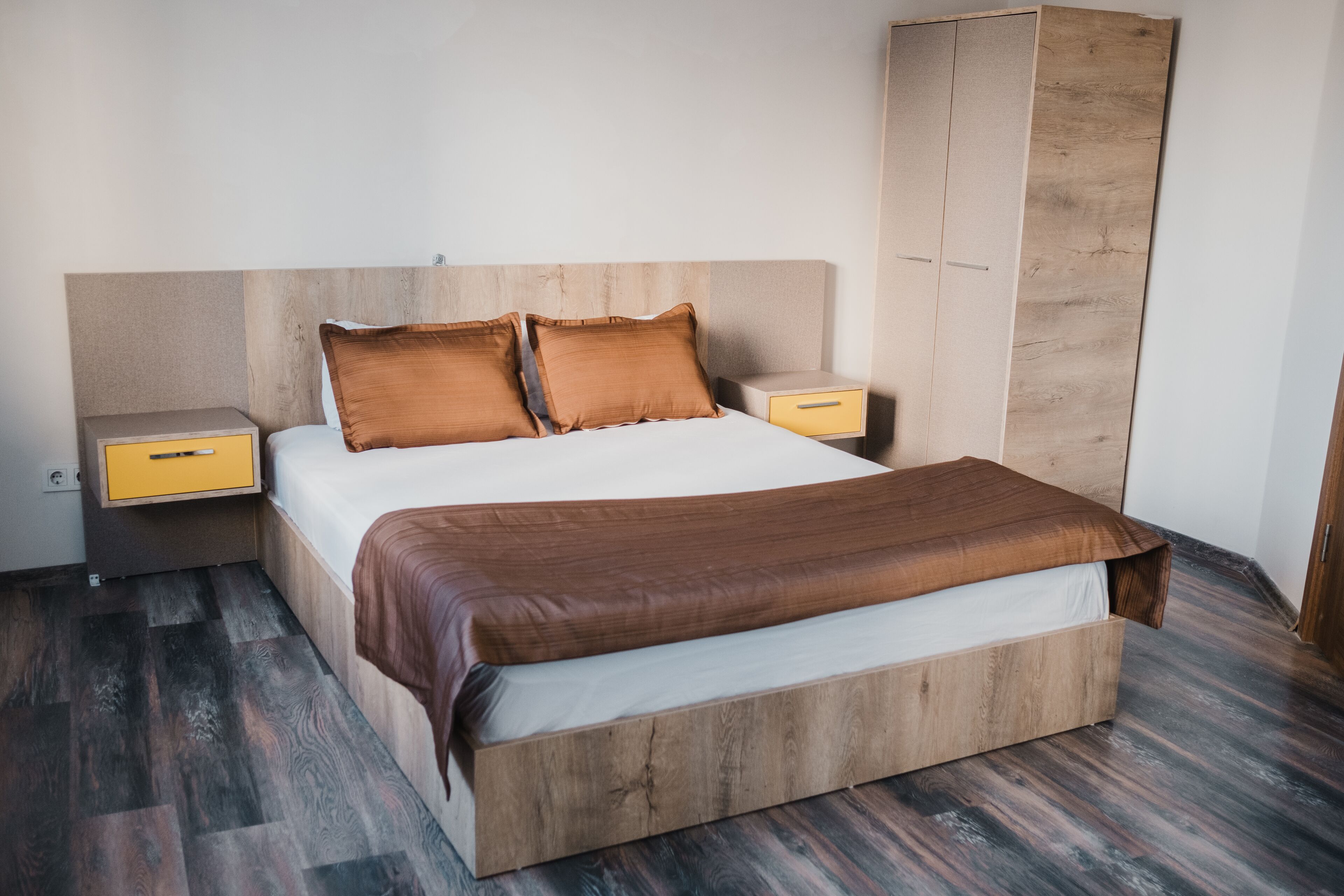 A bedroom with a wooden headboard, a large bed with a brown bedspread, two nightstands with yellow drawers, and a wardrobe.