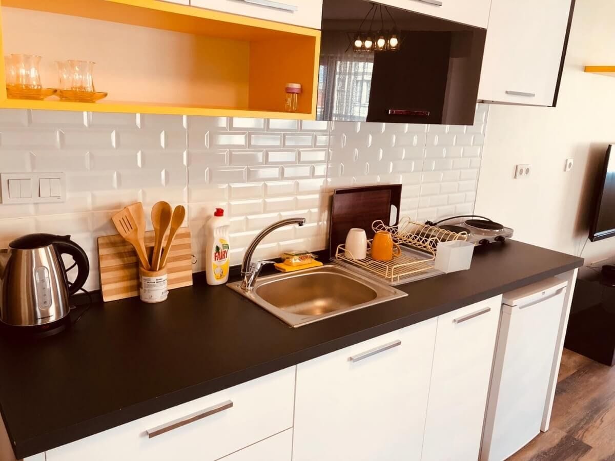 A modern kitchen with a black countertop, white cabinets, and a stainless steel sink. There are wooden utensils in a holder, a kettle, and a dish rack with dishes.
