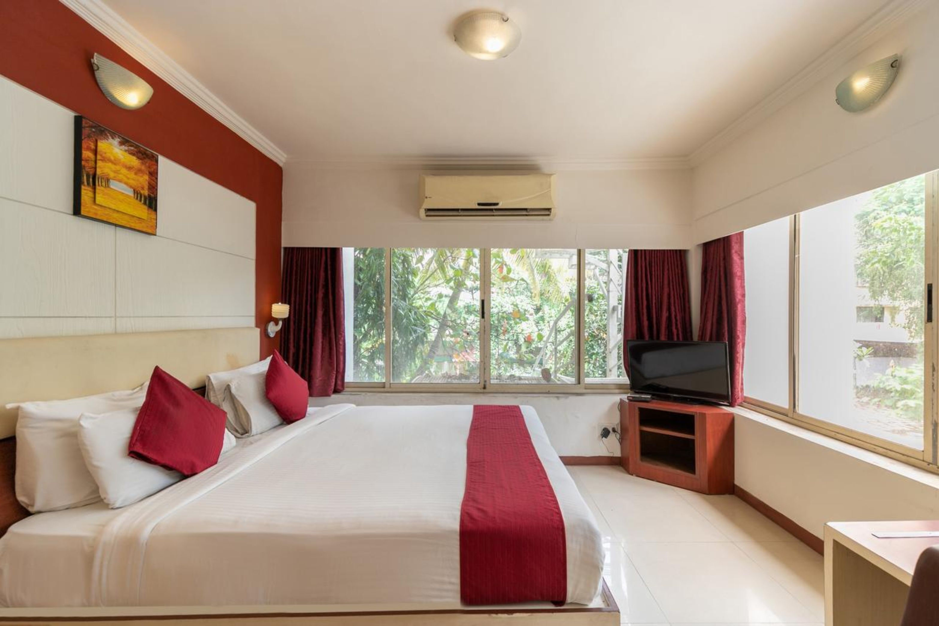 A hotel room with a large bed, a flat-screen TV, and a window with a view of greenery.