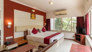 Quarto de hotel com cama, mesa de cabeceira, escrivaninha, cadeira, suporte para TV e janela com cortinas.
