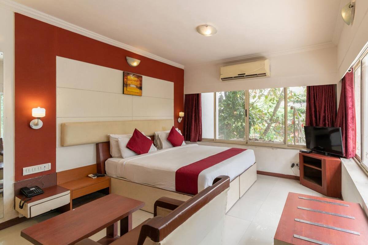 A hotel room with a bed, bedside table, desk, chair, TV stand, and a window with curtains.