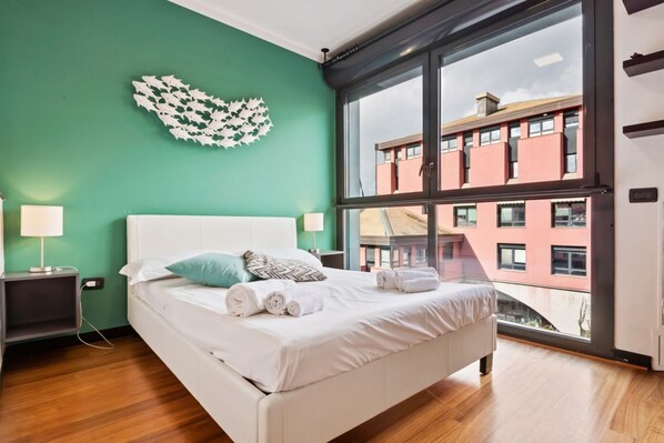 A bedroom with a large window, a bed with white linens, a green wall, and a decorative leaf wall art piece.