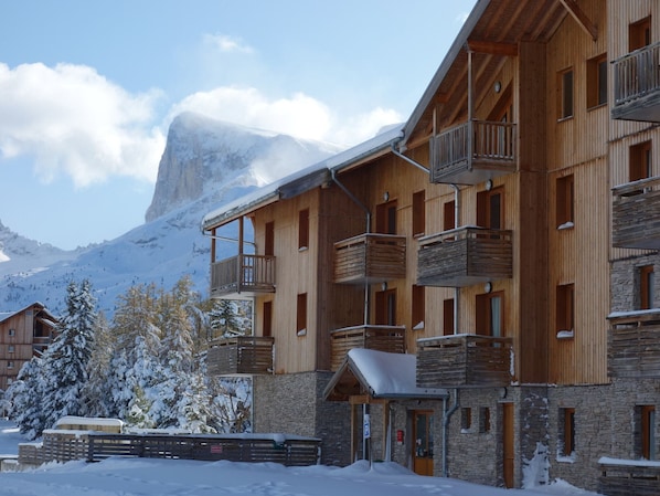 A multi-story wooden building with balconies, a stone facade, and a snowy mountain backdrop. - Charming 3-room chalet for 6 guests in Superdevoluy (Le Dévoluy)
