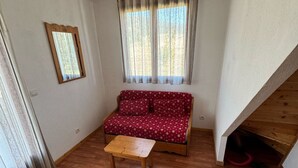A small, cozy room with a wooden floor, a red sofa with floral upholstery, a wooden table, and a window with sheer curtains. - Charming 3-room chalet for 6 guests in Superdevoluy (Le Dévoluy)