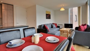Un salon moderne avec un canapé gris, des coussins rouges, une table à manger dressée avec des assiettes et des bougies, et un téléviseur posé sur une table en bois.