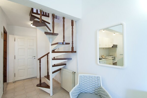 A staircase with wooden railings leading to a white door, a radiator, and a corner with a cushioned chair. - Sofia Studios (Skiathos)