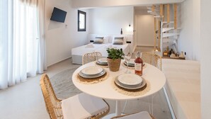 A dining area with a round table set for four, surrounded by wicker chairs with white cushions. A woven basket with a plant and a bottle of wine are on the table. - Sofia Studios (Skiathos)
