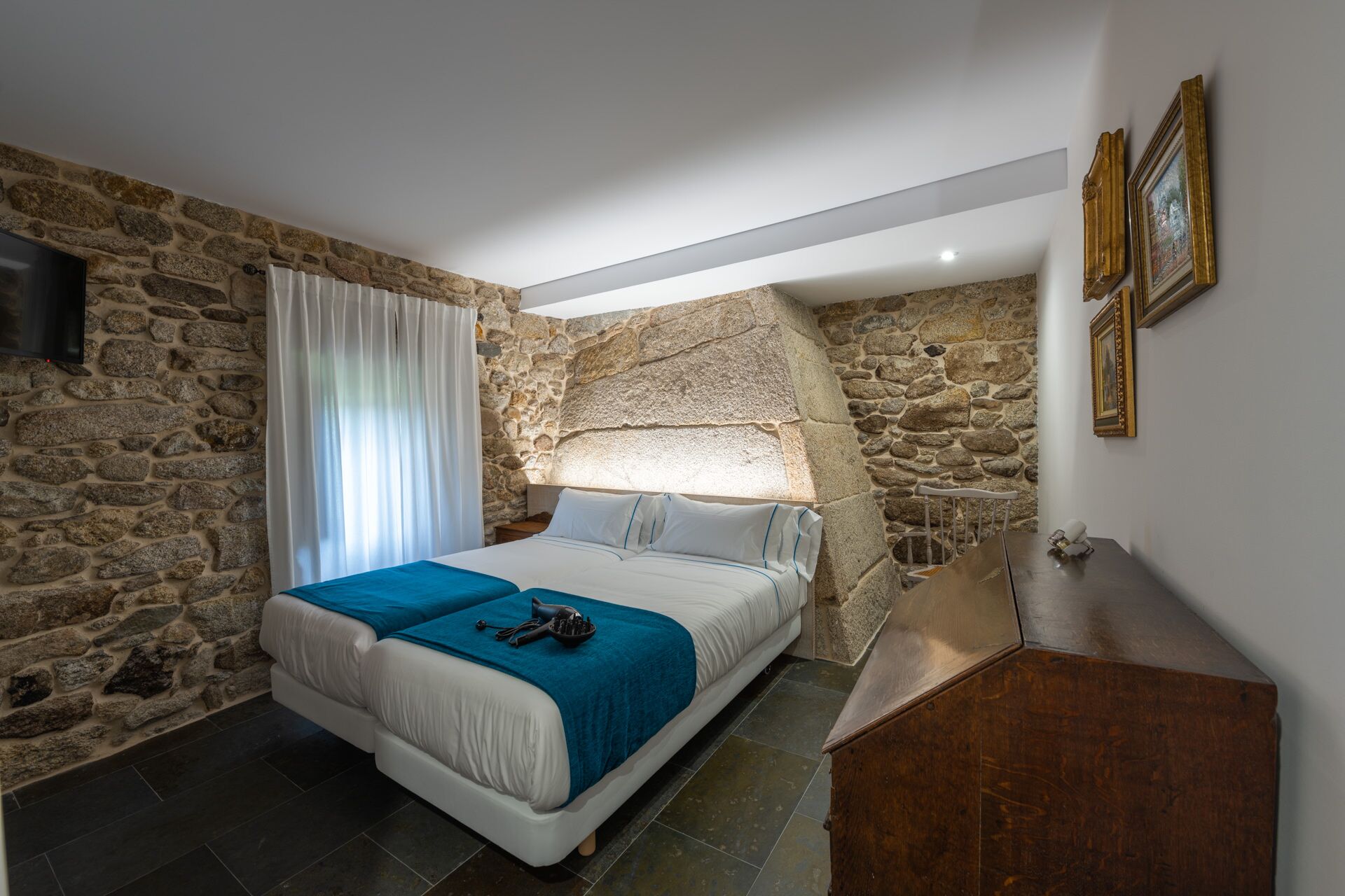 Une chambre avec un mur en pierre, un lit recouvert d’une couverture bleue, une commode en bois et un téléviseur fixé au mur.