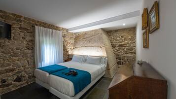 Un dormitorio con pared de piedra, una cama con manta azul, un aparador de madera y un televisor montado en la pared.