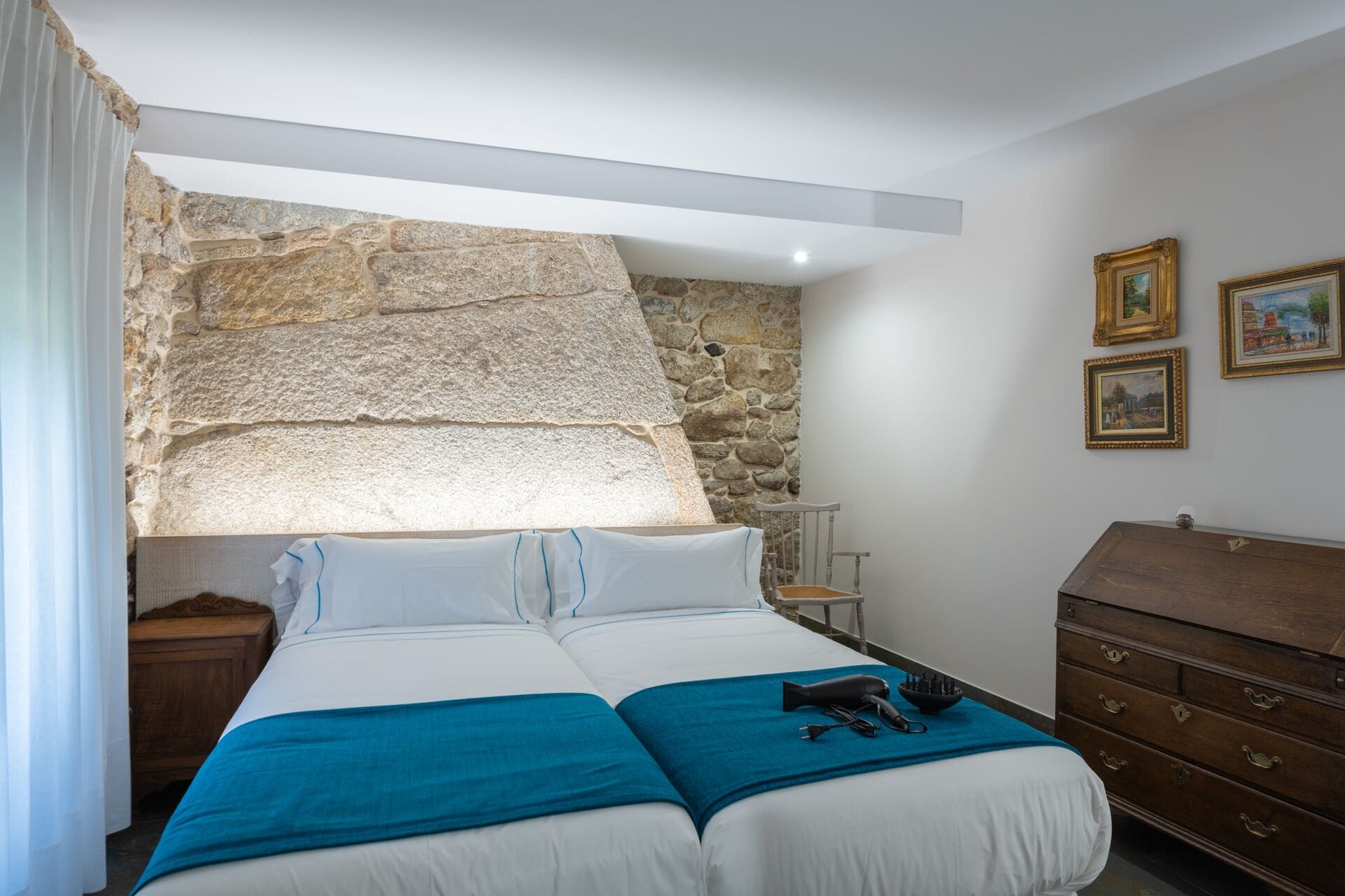 A bedroom with a stone wall, a bed with white bedding, a blue throw, and a wooden dresser.