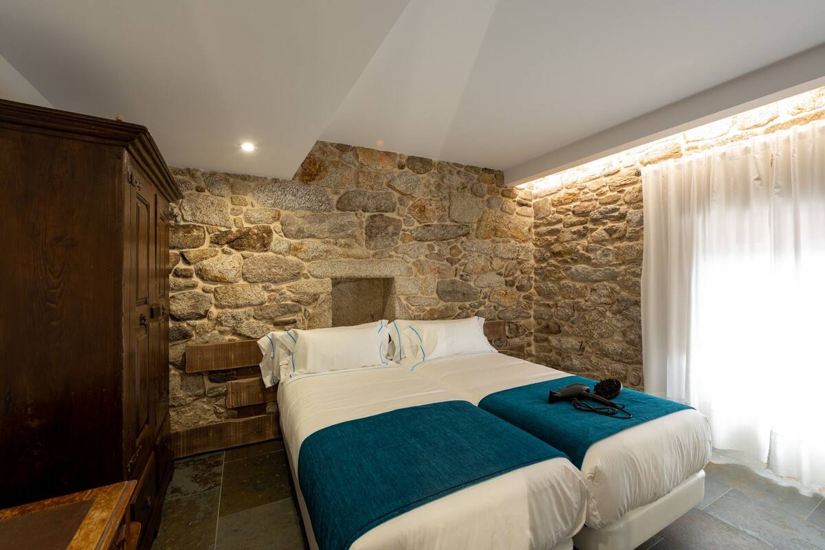 Quarto com parede de pedra, guarda-roupa de madeira, cama com duas almofadas e janela com cortinas.