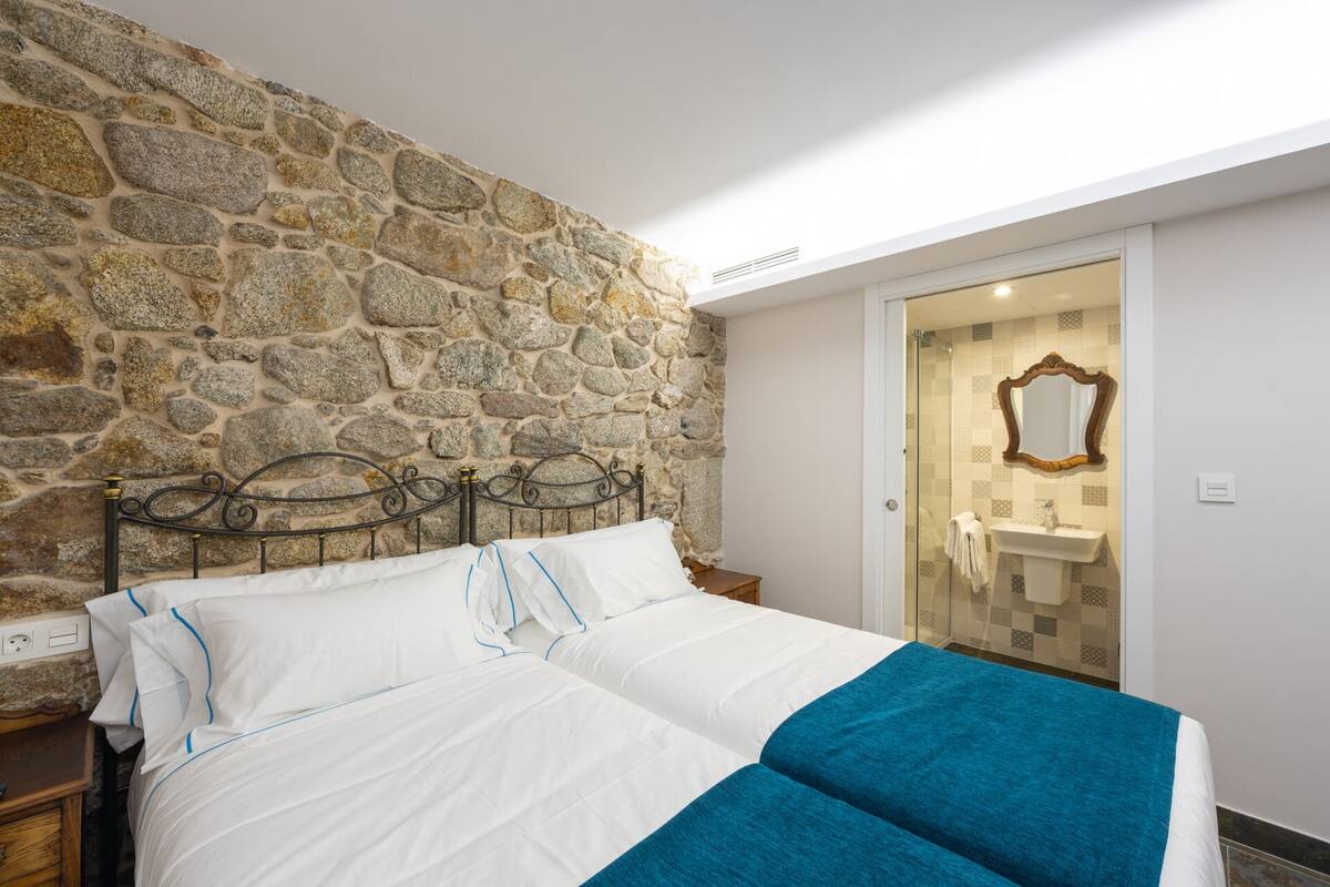 Quarto com parede de pedra, cama com roupa de cama branca e um toque de azul, mesa de cabeceira de madeira e banheiro visível por uma porta de vidro.