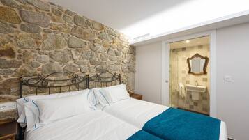 Ein Schlafzimmer mit einer Steinswand, einem Bett mit weißer Bettwäsche und blauem Akzent, einem Nachttisch aus Holz und einem durch eine Glastür sichtbaren Badezimmer.