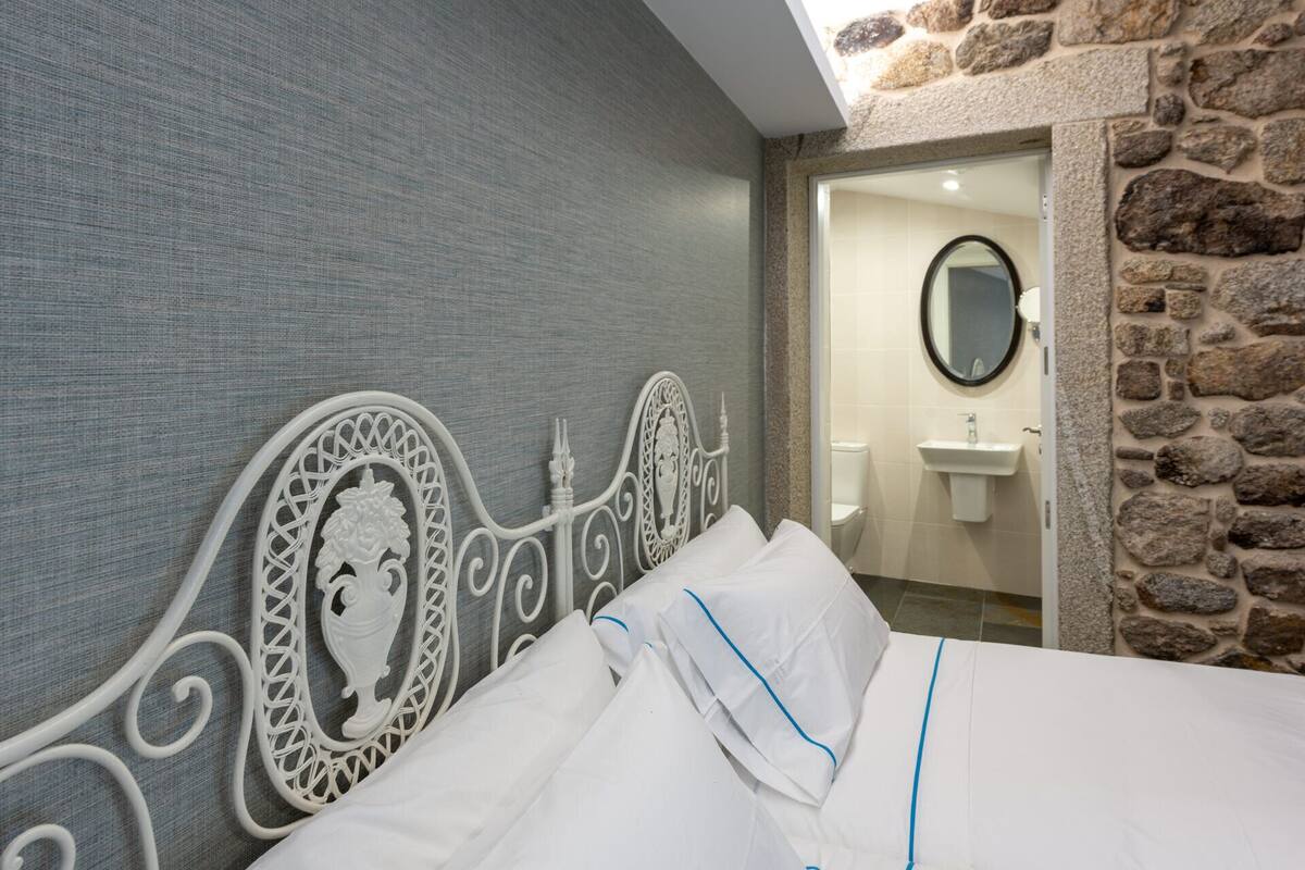 Cama bem arrumada com lençóis brancos e detalhes azuis, cabeceira de metal decorativa e parede de pedra adjacente a um banheiro com pia e espelho.