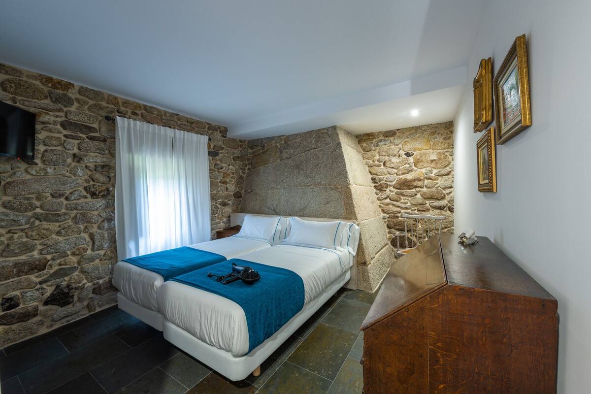 Quarto com parede de pedra, cama com roupa de cama azul e branca, penteadeira de madeira e um quadro na parede.