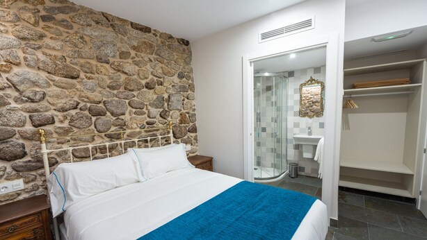 Ein Schlafzimmer mit Steinwand, einem Bett mit weißer Bettwäsche und einer blauen Tagesdecke, einem hölzernen Nachttisch und einem Kleiderschrank mit Spiegel.