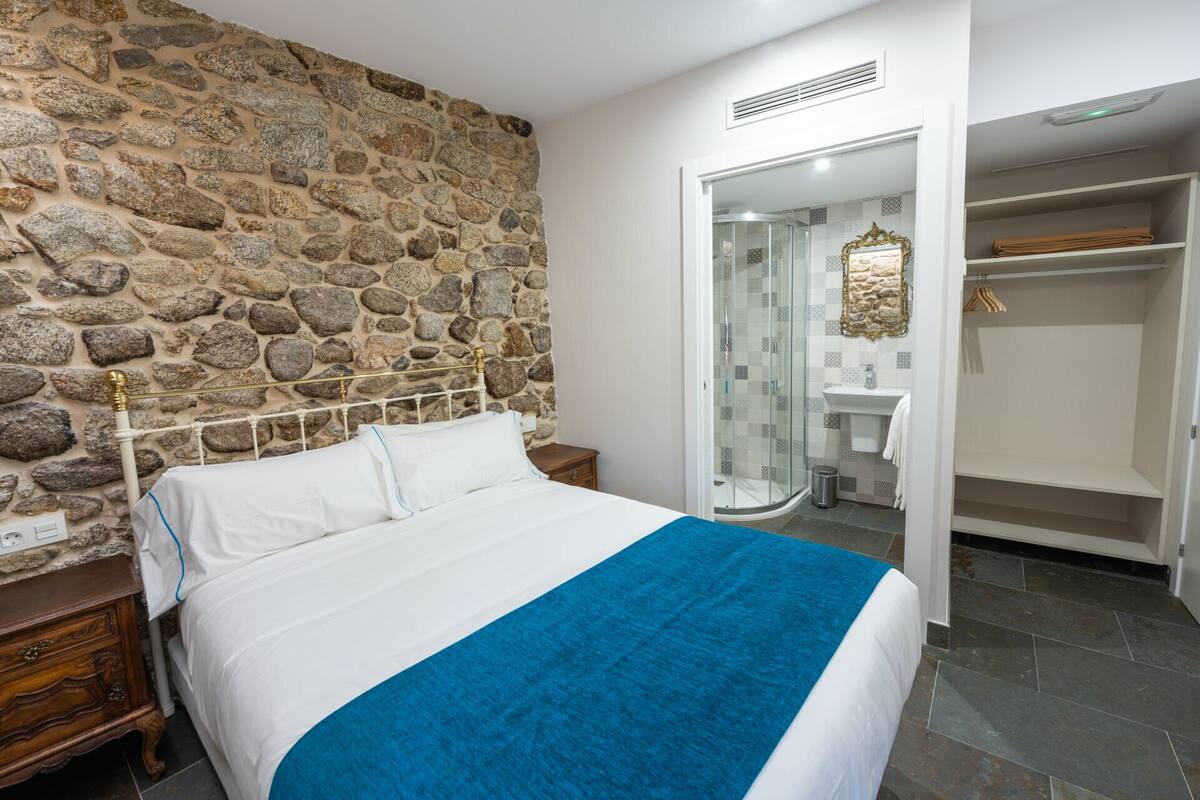 Quarto com parede de pedra, cama com roupa de cama branca e manta azul, mesa de cabeceira de madeira e armário com espelho.