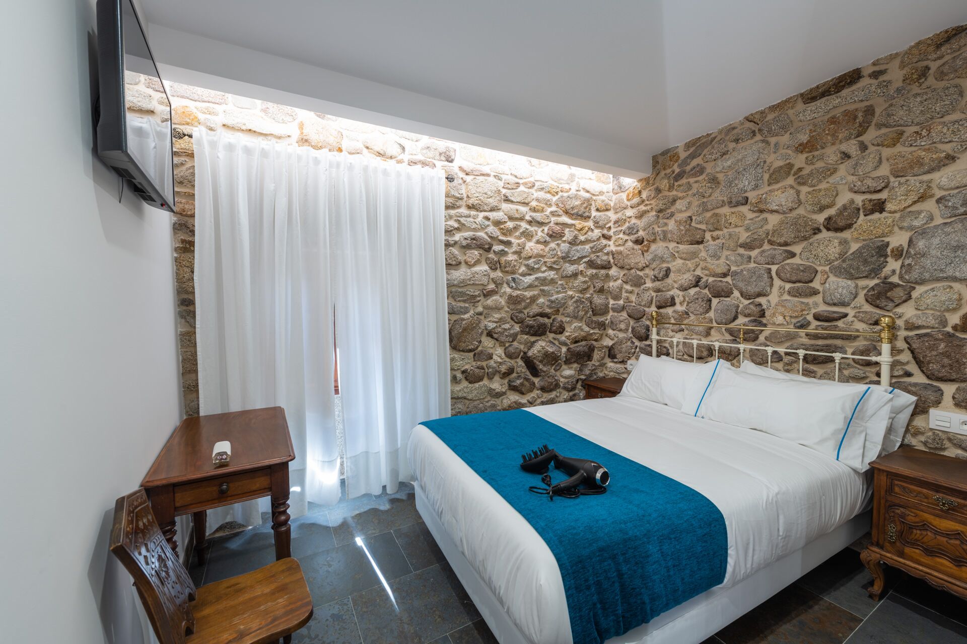 Un dormitorio con pared de piedra, cama con ropa blanca y un plaid azul, mesita de noche de madera y televisor montado en la pared.