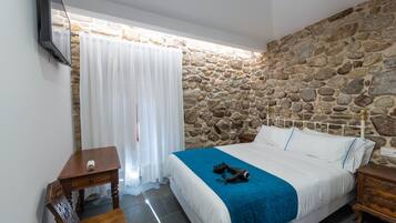 Un dormitorio con pared de piedra, cama con ropa blanca y un plaid azul, mesita de noche de madera y televisor montado en la pared.