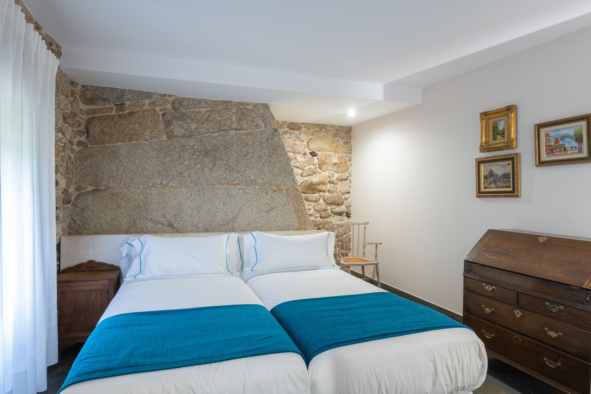 Une chambre avec un mur en pierre, un lit avec une literie bleue, une commode en bois et deux tableaux encadrés accrochés au mur.