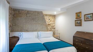 A bedroom with a stone wall, a bed with blue bedding, a wooden dresser, and two framed pictures on the wall.