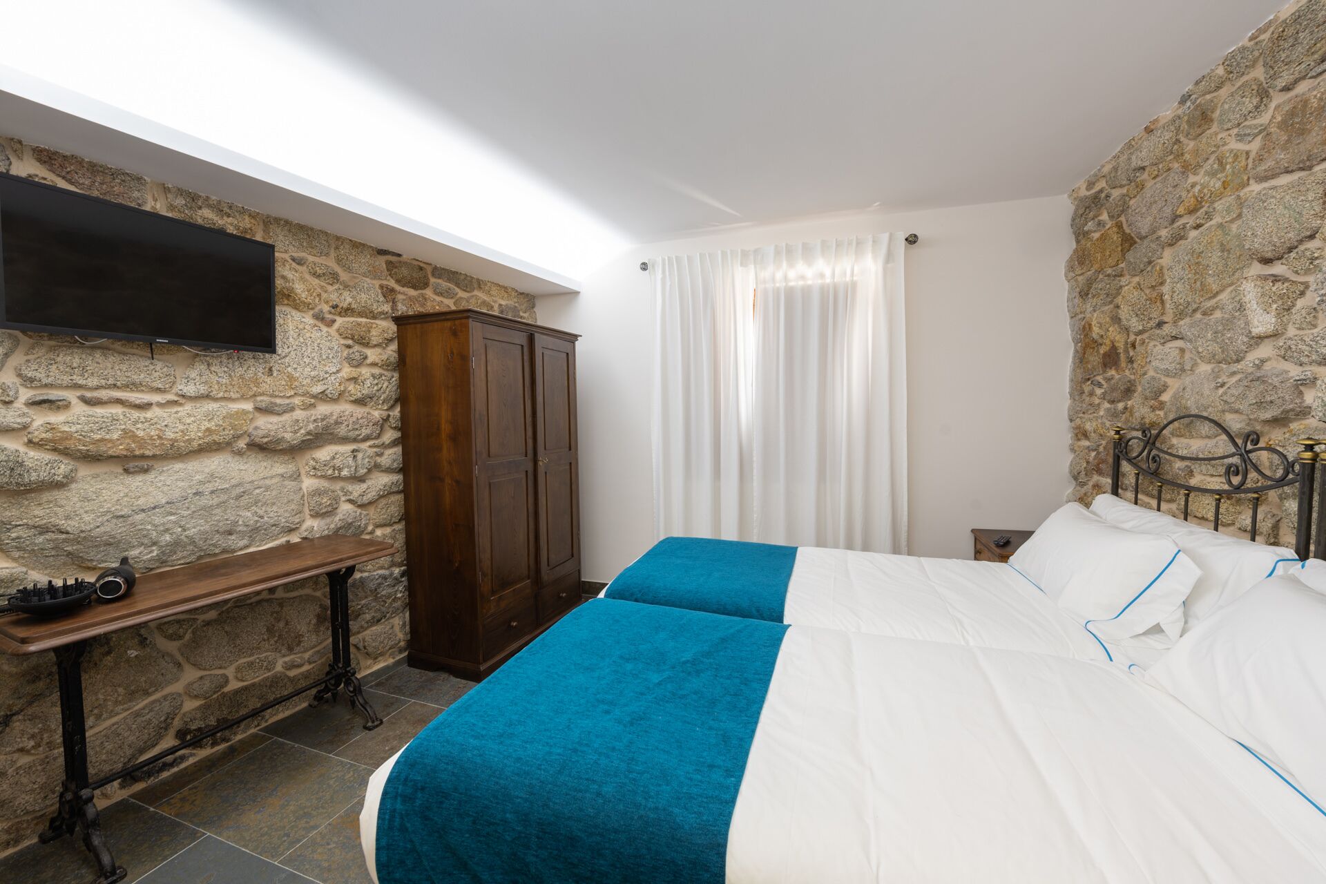 A bedroom with a stone wall, a wooden wardrobe, a bed with blue and white bedding, and a flat-screen TV mounted on the wall.