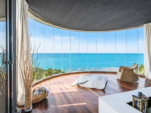Une pièce moderne avec un plancher en bois aux lignes courbes, de grandes fenêtres donnant sur la mer, un fauteuil en osier et un canapé blanc.