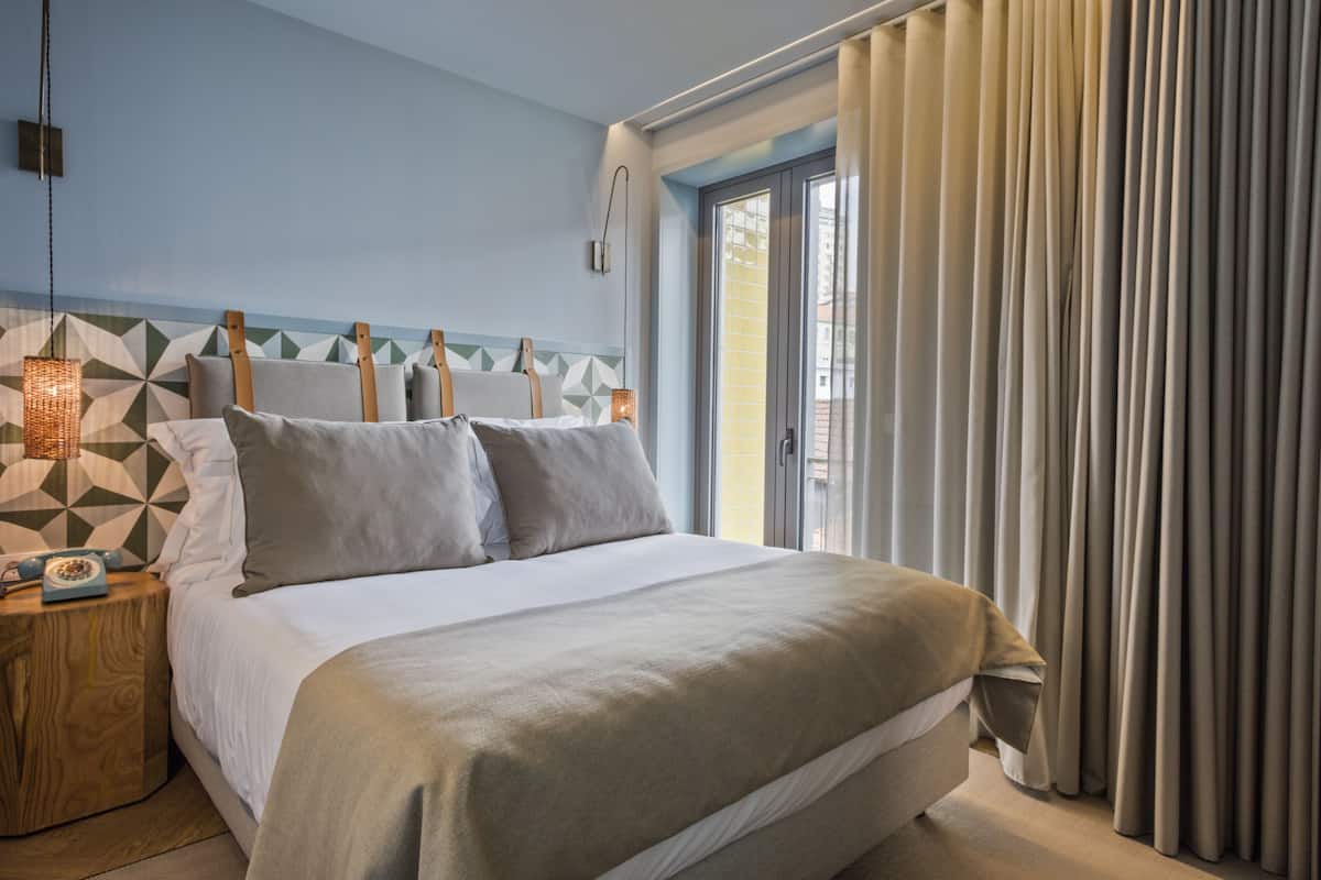 Een moderne slaapkamer met een groot bed, een hoofdbord met een geometrisch patroon, een bedlampje en uitzicht op het omliggende landschap door een raam met gordijnen.