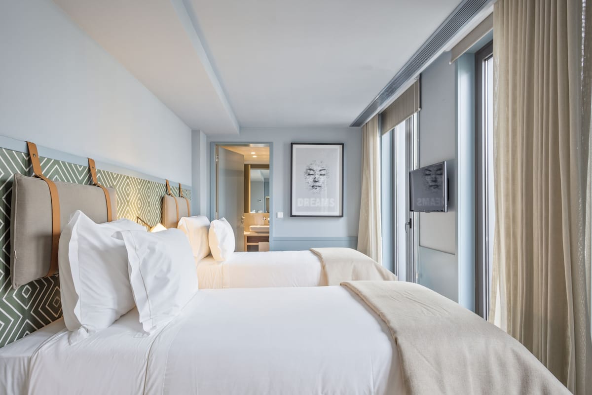 Een hotelkamer met twee bedden, een groot raam met gordijnen, een kunstwerk in een lijst aan de muur, en een deur naar een badkamer.