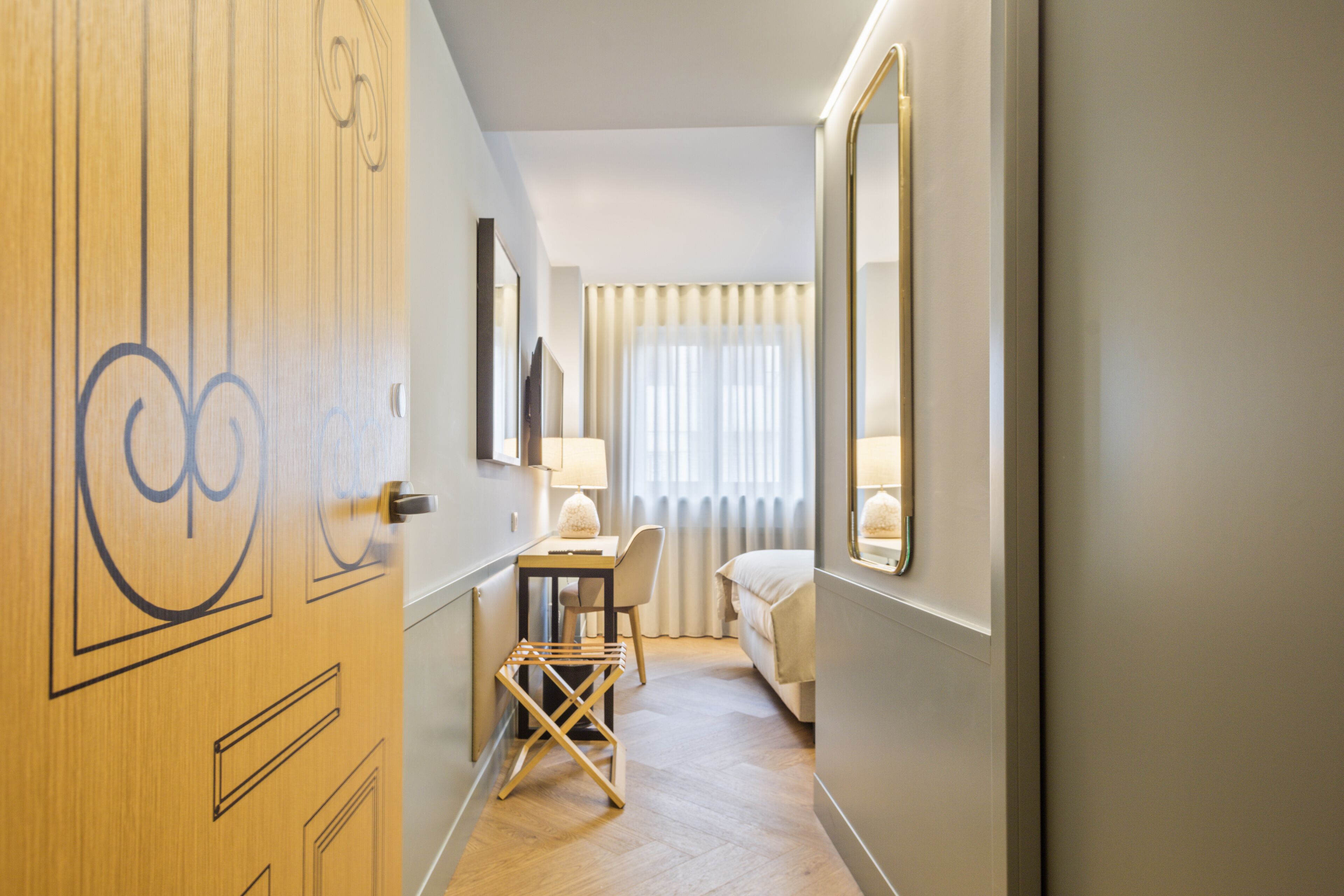 A hotel room with a wooden door, a desk with a chair, a bed, a window with curtains, and a mirror on the wall.